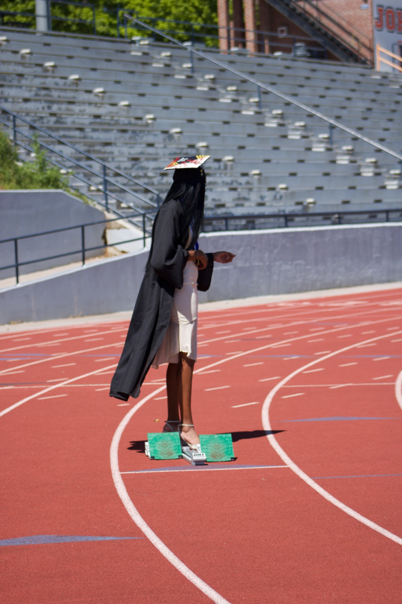 Doyle stands on a block on the track in her graduation attire, blending both of her worlds of star athlete and student scholar.