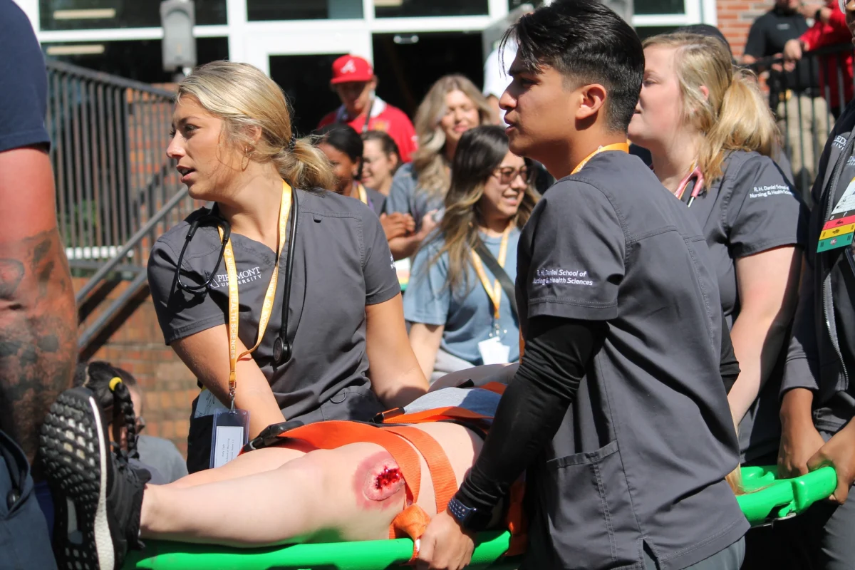 Nursing students carry out a wounded student with a massive gash on her leg. Using a spine-board to carry her out the building and down the stairs.